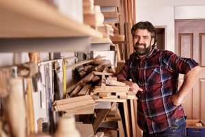 Smiling carpenter and small business owner, looking at the camera, while standing confidently in his workshop with shelves of wooden planks